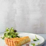 A slice of a creamy onion tart, with a flaky crust and tossed mixed salad on a white plate