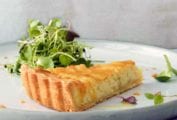A slice of a creamy onion tart, with a flaky crust and tossed mixed salad on a white plate