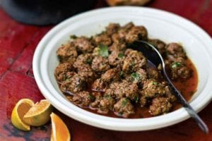 A white bowl filled with Moroccan meatballs with a spoon resting inside and some small orange wedges on the side.