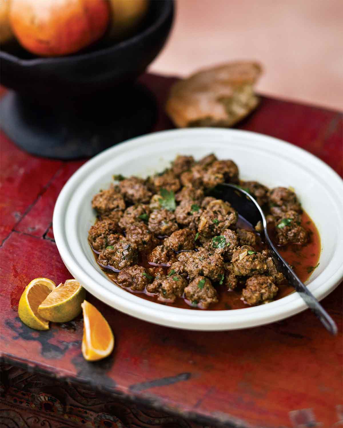 A white bowl filled with Moroccan meatballs with a spoon resting inside and some small orange wedges on the side.