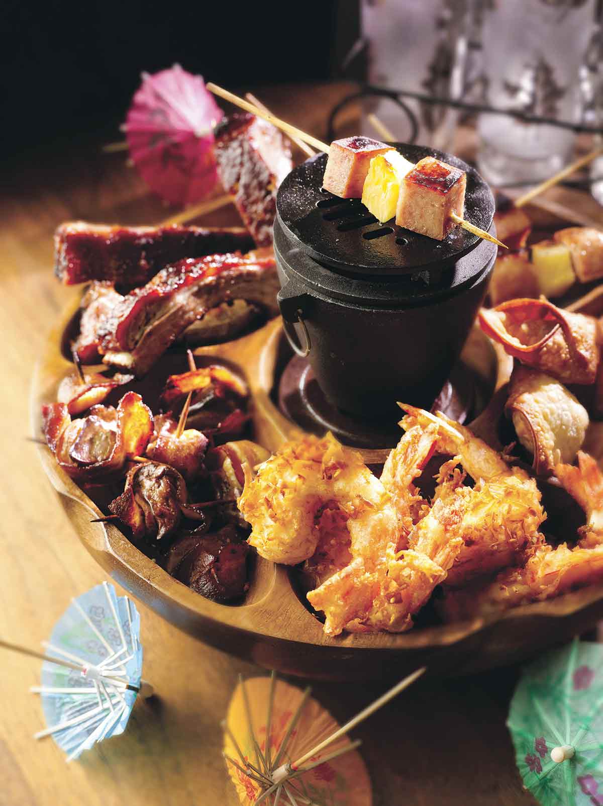 A wooden pupu platter filled with coconut shrimp, spam and pineapple skewers, and ribs.