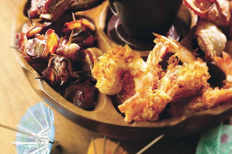 A wooden pupu platter filled with coconut shrimp, spam and pineapple skewers, and ribs.
