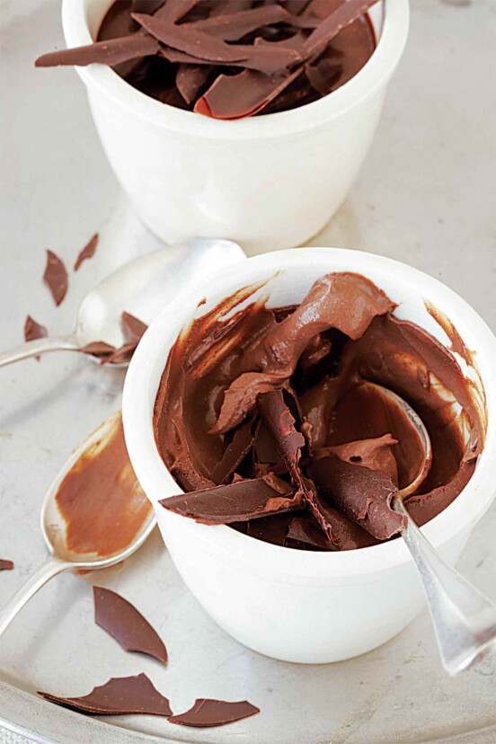 A chocolate pot de crème topped with chocolate shavings in a white cup with two spoons resting beside.