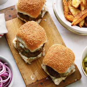 Three lamb burgers with chunky potato wedges and sliced pickles in separate bowls.