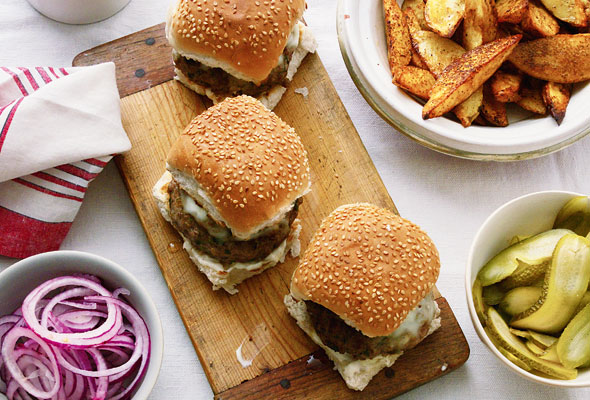 Three lamb burgers with chunky potato wedges and sliced pickles in separate bowls.