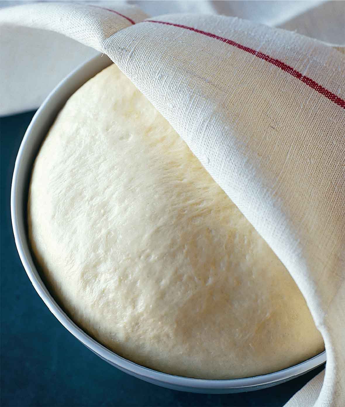 A ball of rising semolina pizza dough in a round metal tin, partially covered by a kitchen towel.