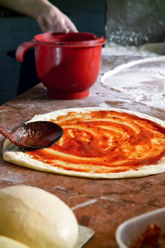 A wooden spoon spreading simple pizza sauce on a round of pizza dough.