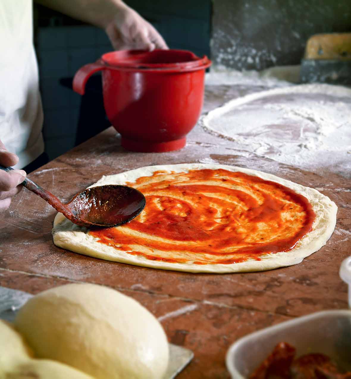 A wooden spoon spreading simple pizza sauce on a round of pizza dough.