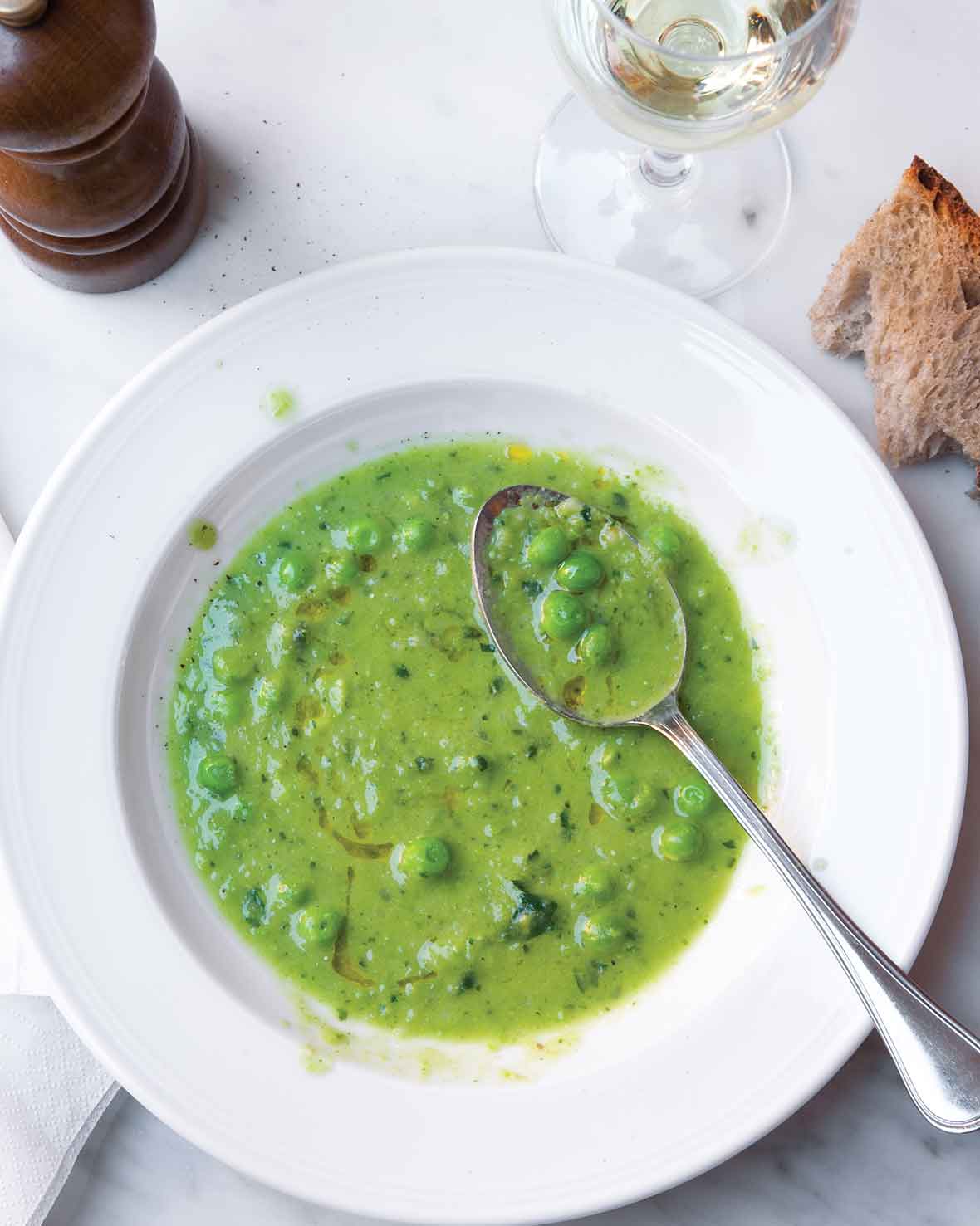 A bowl of pureed pea soup with mint with a spoon resting in the bowl and a glass of wine, piece of bread, and pepper mill beside it.