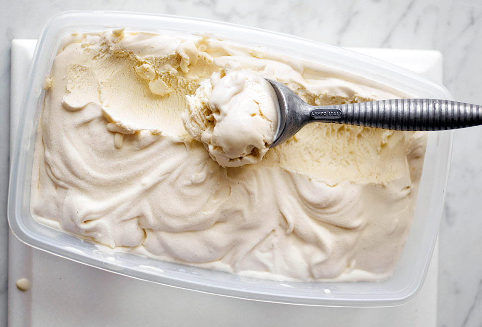 A plastic container filled with vanilla bean ice cream with a metal ice cream scoop resting inside.