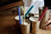 Four cereal milk white Russian cocktails in copper cups, being poured from a pitcher, with colored straws, with a person's hand holding a shaker in the background.
