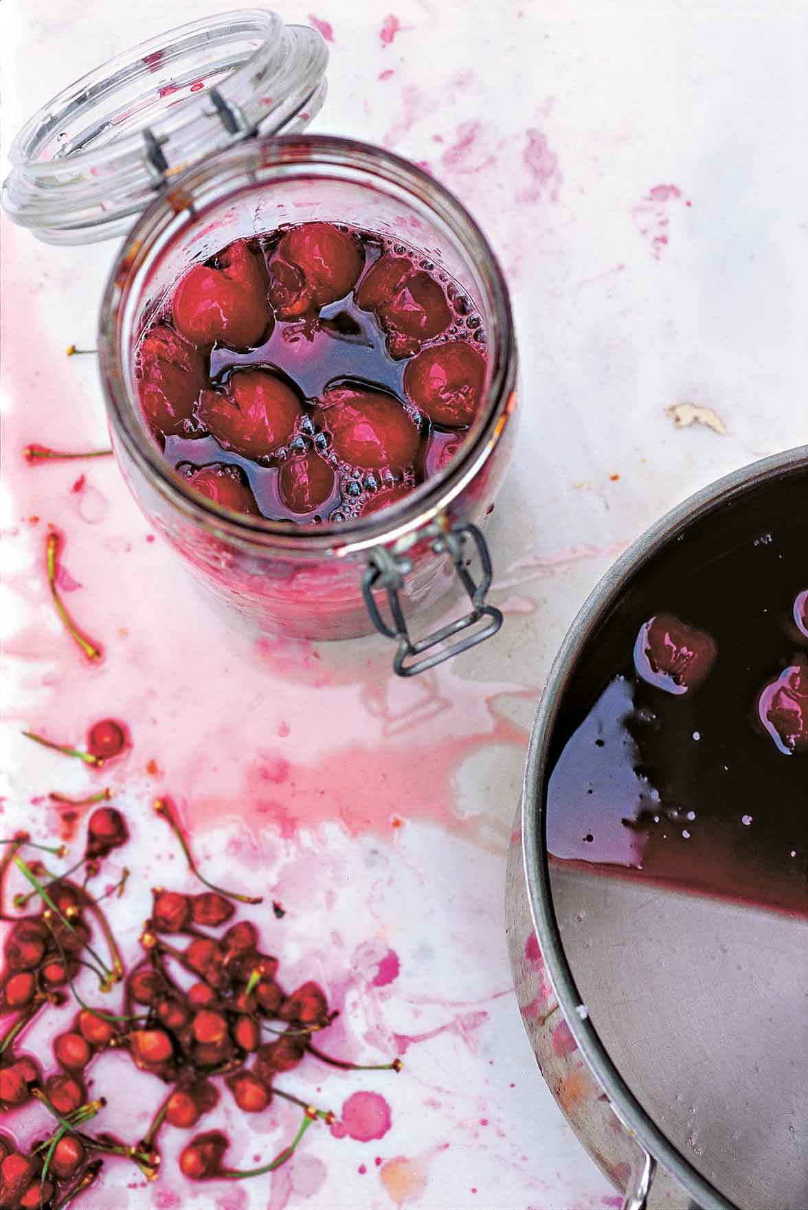 A canning jar filled with cherry liqueur and whole cherries, with a pot of liqueur and many cherry stones beside it.