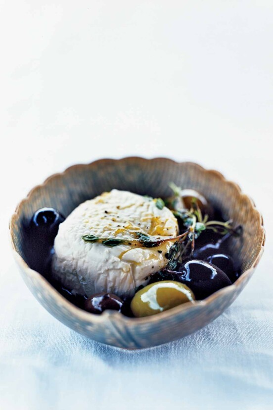 A decorative bowl filled with a sphere of goat cheese and a few olives, topped with a thyme sprig.
