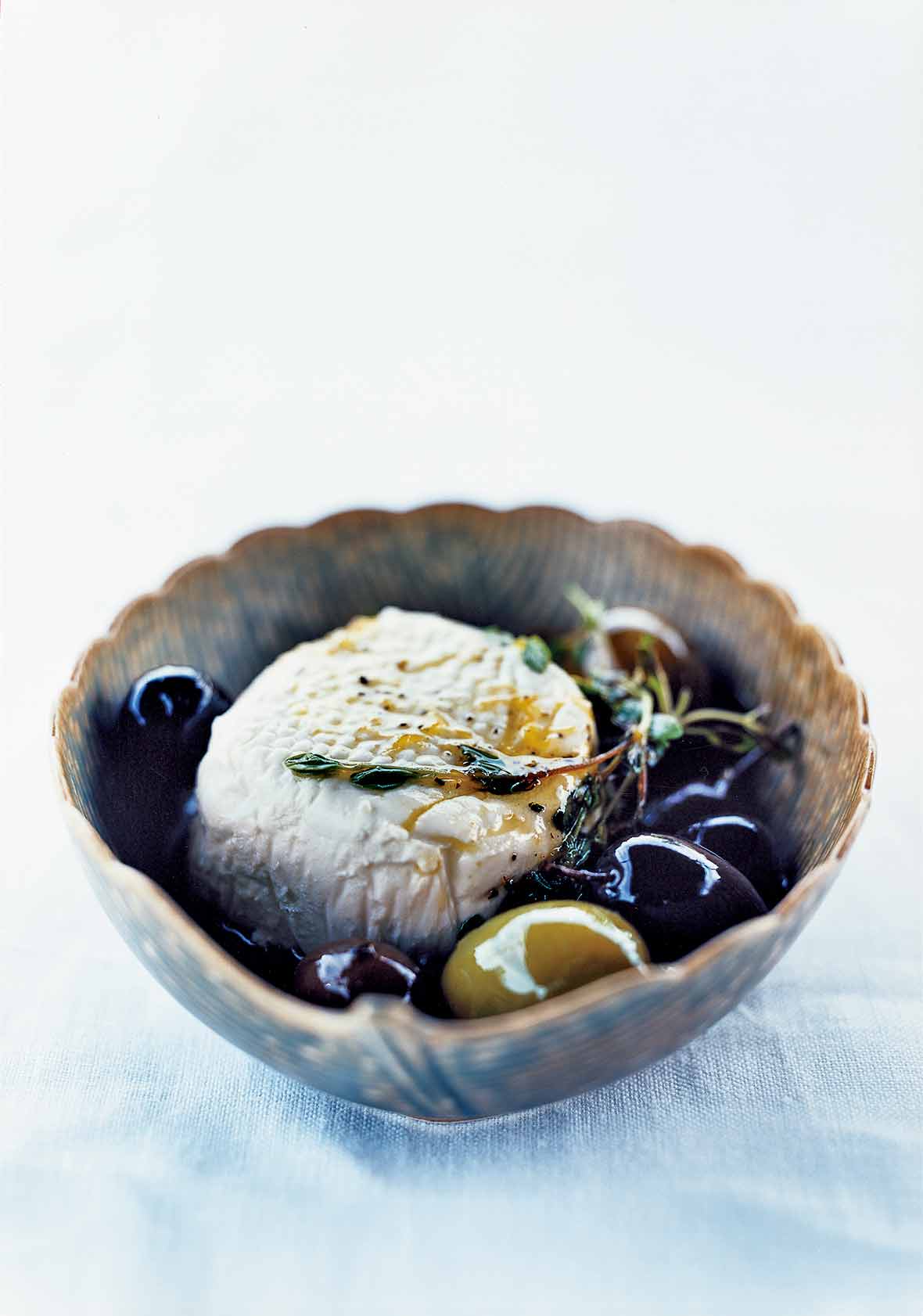 A decorative bowl filled with a sphere of goat cheese and a few olives, topped with a thyme sprig.