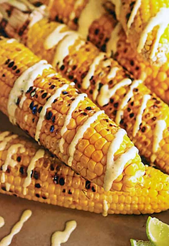 Six ears of grilled Indian corn drizzled with curry yogurt and lime wedges on the side.
