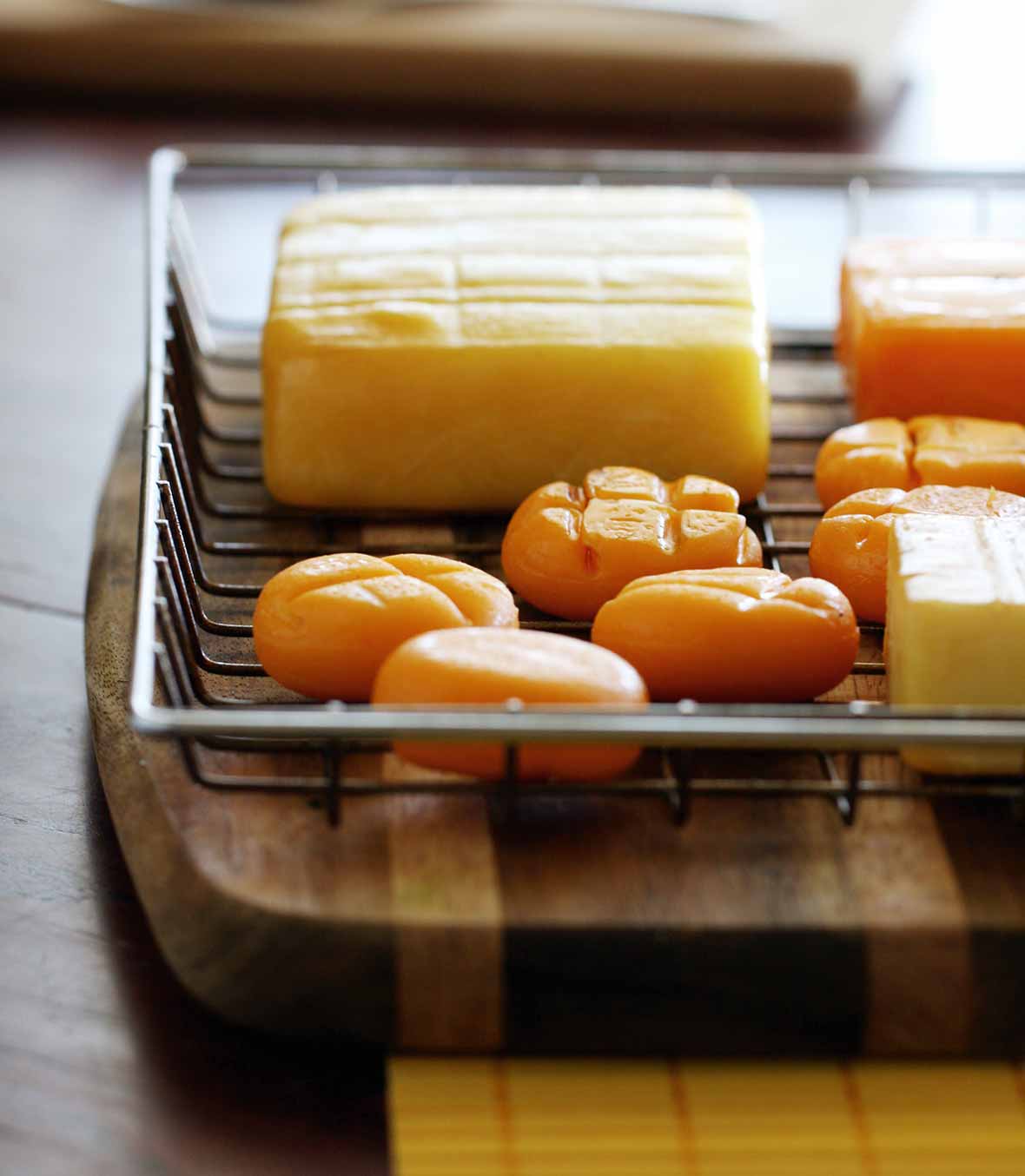 A wire basket on a wood cutting board containing several types of cheese, including smoked cheddar cheese.