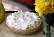 A banana cream pie, topped with pillowy meringue in a glass pie dish with a vase of daffodils beside it.