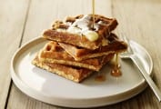 Four cornmeal and oat waffles stacked on a white plate, covered with butter and having syrup poured on them.