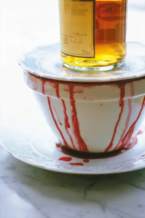 Wine-soaked autumn pudding in a white bowl with wine dripping down the side topped with a white plate and a bottle of whiskey.