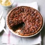 A chocolate pecan pie in a ceramic pie plate with one slice missing on a linen napkin.
