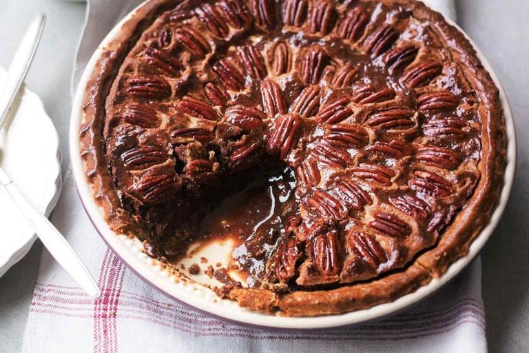 A chocolate pecan pie in a ceramic pie plate with one slice missing on a linen napkin.