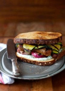 A grilled sausage sandwich with strips of grilled zucchini, red onion, and a herb mayo on a silver plate with a knife resting beside it.