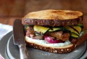 A grilled sausage sandwich with strips of grilled zucchini, red onion, and a herb mayo on a silver plate with a knife resting beside it.