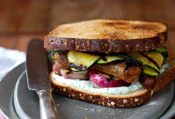 A grilled sausage sandwich with strips of grilled zucchini, red onion, and a herb mayo on a silver plate with a knife resting beside it.