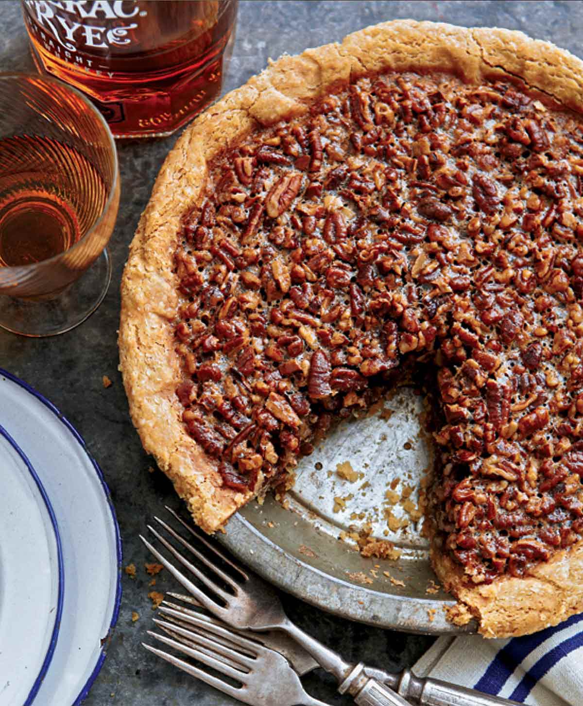 A maple pecan pie in a metal pie plate with one slice missing and forks, plates, and a bottle of rye whiskey beside the pie.