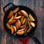 A cast-iron skillet filled with roasted grilled potatoes and two sprigs of rosemary on a wooden table.