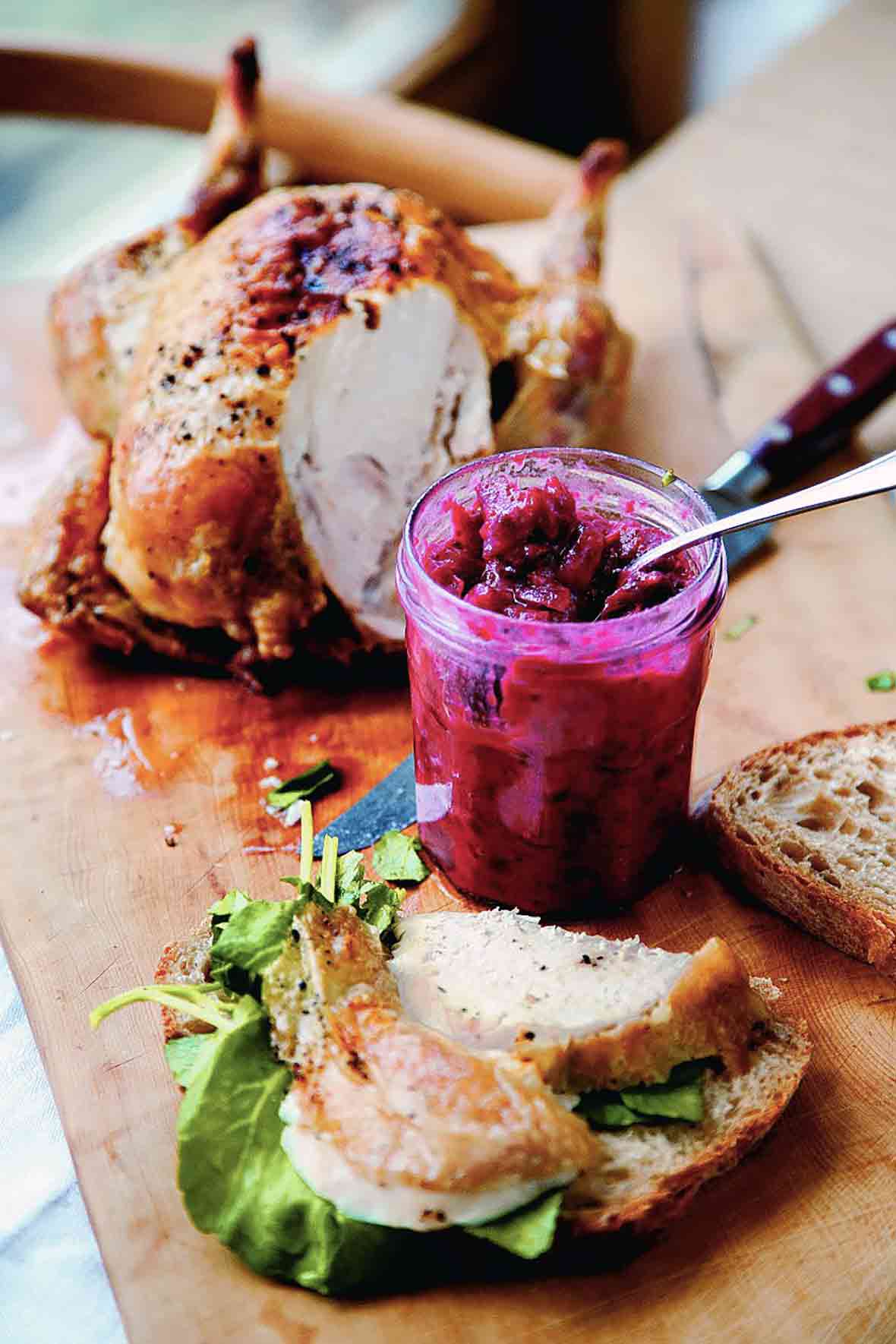 An open jar of cranberry chutney on a cutting board next to a partially carved chicken and a sandwich being assembled.