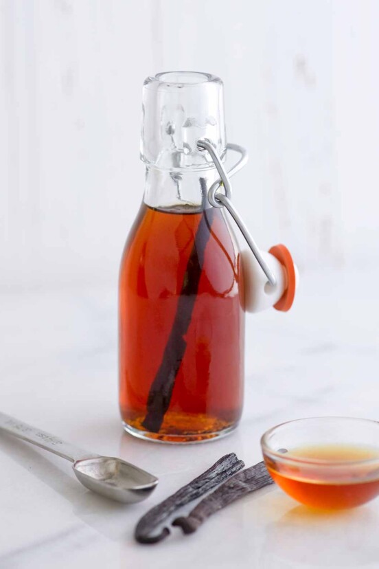 A small bottle of homemade vanilla extract, a measuring spoon, two vanilla beans, and a small bowl of amber liquid.