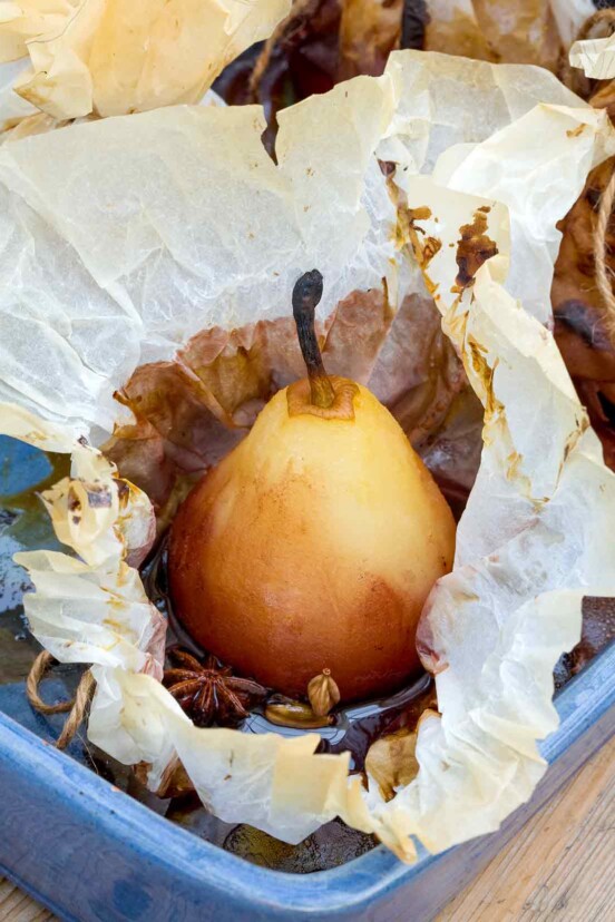 A whole peeled pear in an opened piece of parchment with star anise and cardamom in a blue baking dish.