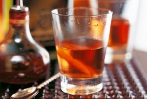 A Sazerac cocktail garnished with a twist, sitting on a bar beside a spoon and a bottle of rye whiskey.