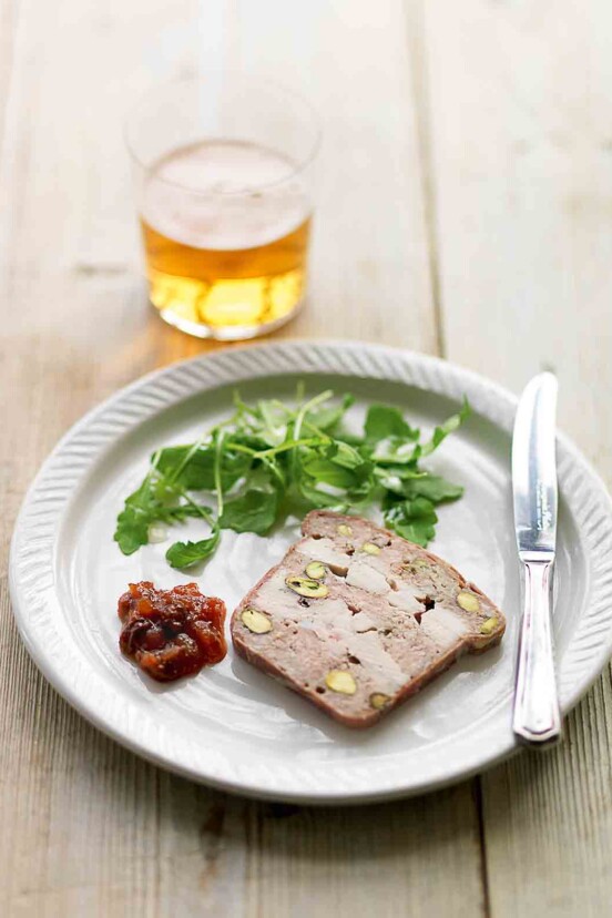 A white plate topped with a slice of chicken, pork, and pistachio terrine, a dollop of chutney, some arugula leaves and a knife.