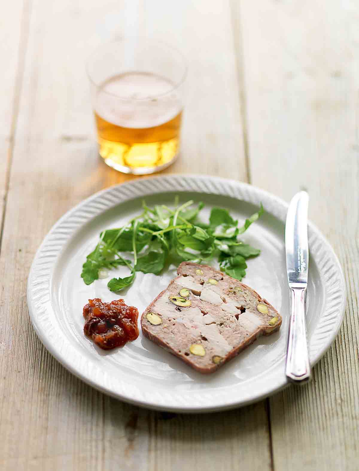 A white plate topped with a slice of chicken, pork, and pistachio terrine, a dollop of chutney, some arugula leaves and a knife.