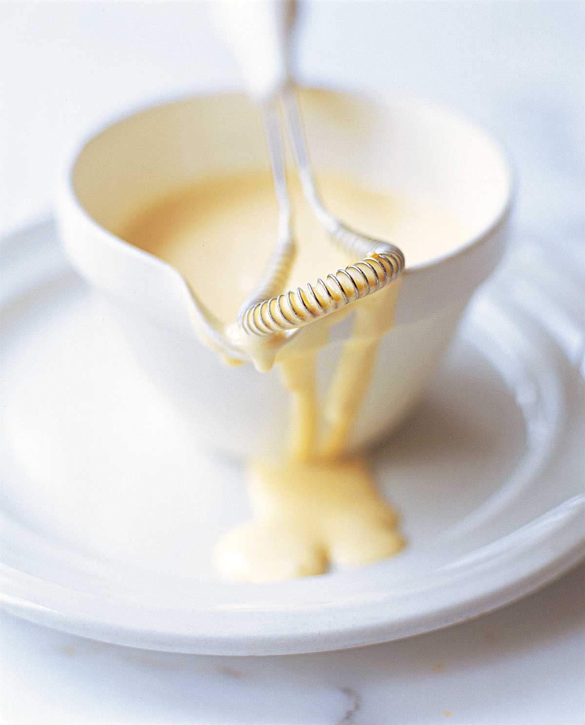 A dish of classic Hollandaise sauce on a white plate with a whisk resting on the dish.