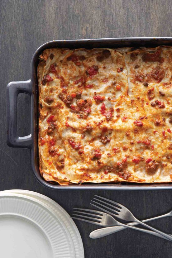 A metal casserole dish filled with a cooked lasagna Bolognese, with three forks and a stack of plates beside it.