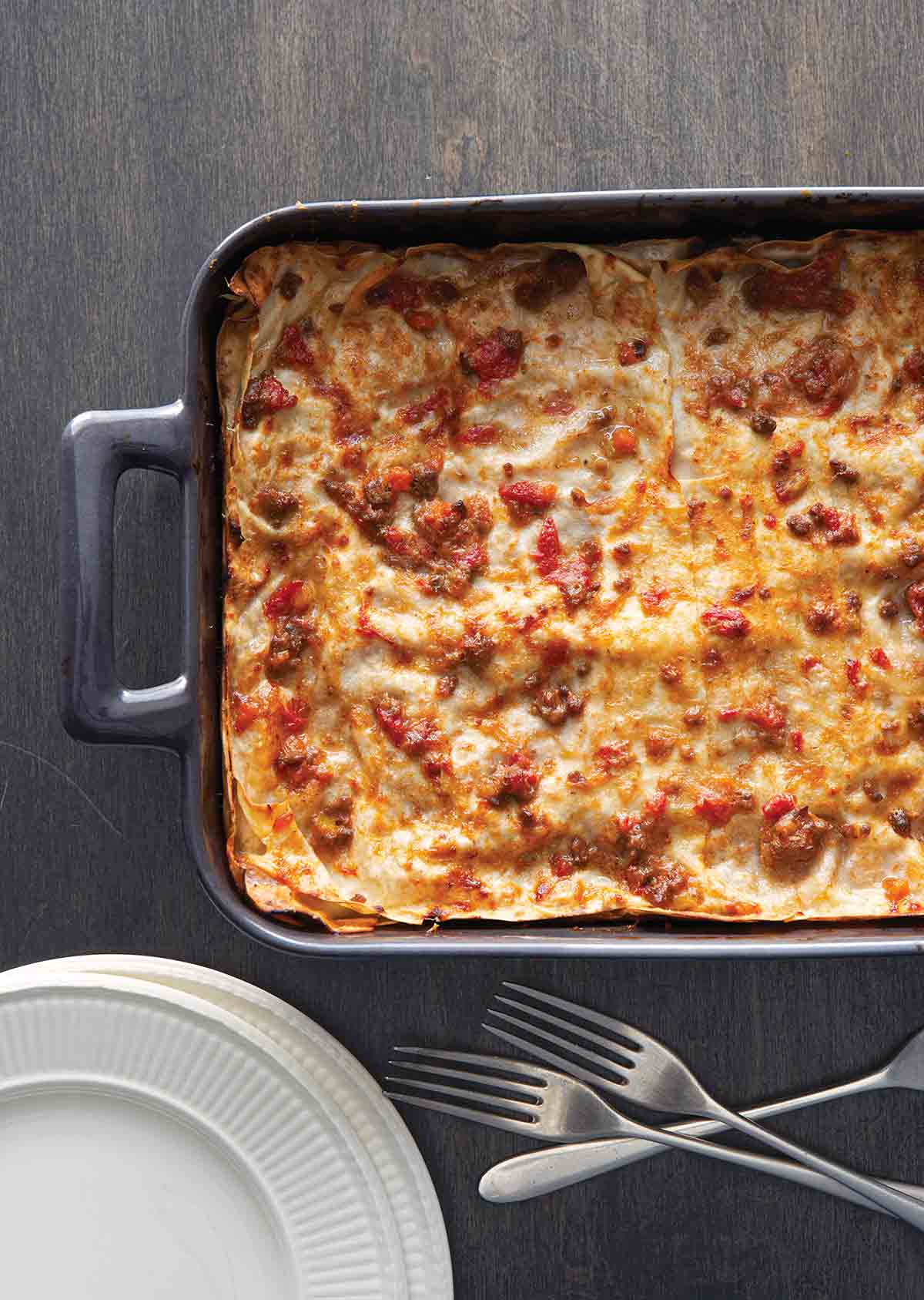 A metal casserole dish filled with a cooked lasagna Bolognese, with three forks and a stack of plates beside it.