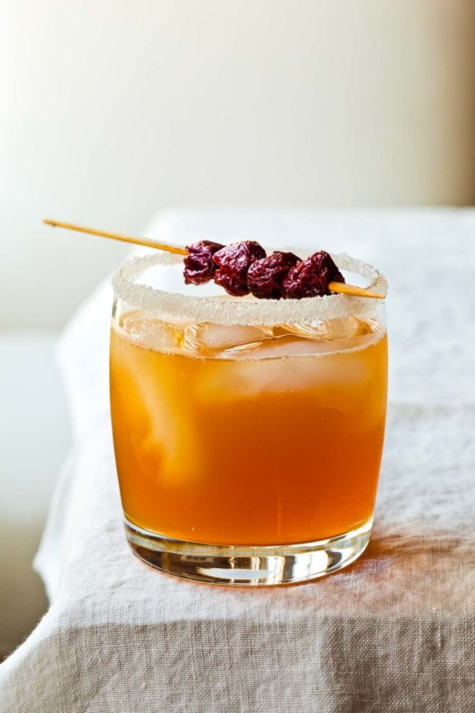 A sidecar cocktail, ice cubes and a dried cherry garnish, in a highball glass with a sugared rim.