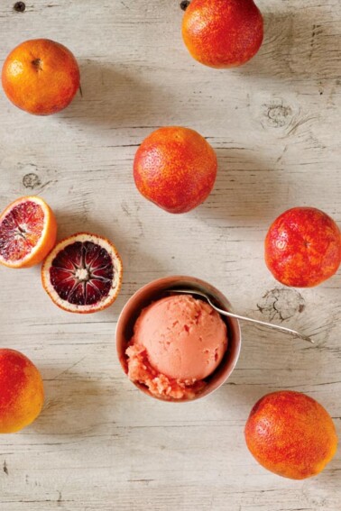 A bowl with a scoop of blood orange sorbet and a spoon in it, surrounded by several blood oranges.