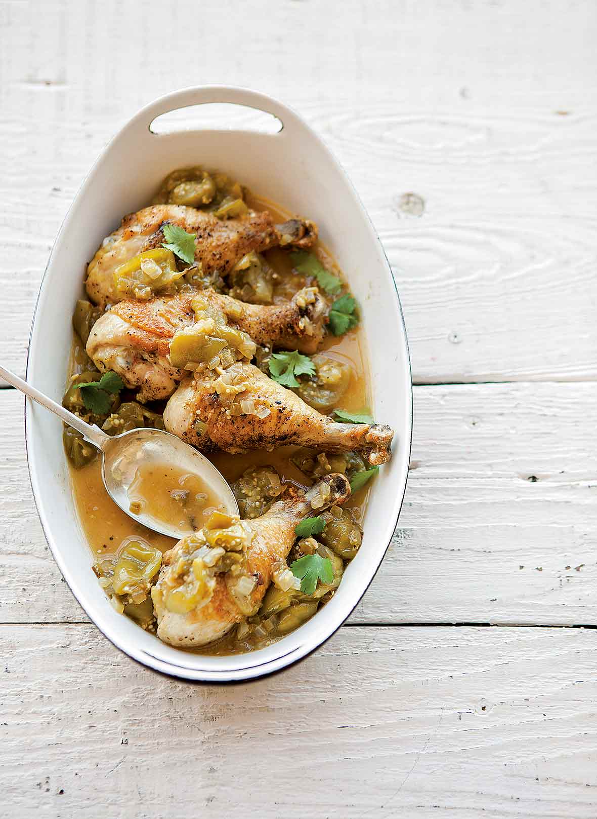 White oval dish with four chicken drumsticks, chopped green tomatillos, cilantro, onion, sliced chiles.