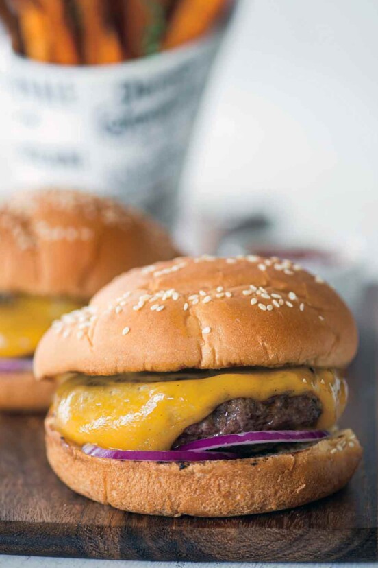 Two classic cheeseburgers with red onion and pickles on sesame buns on a wooden serving board.