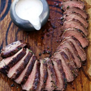 A wooden cutting board topped with sliced loin of grilled venison with horseradish cream sauce on the side.