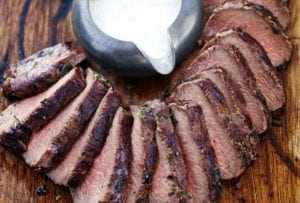 A wooden cutting board topped with sliced loin of grilled venison with horseradish cream sauce on the side.