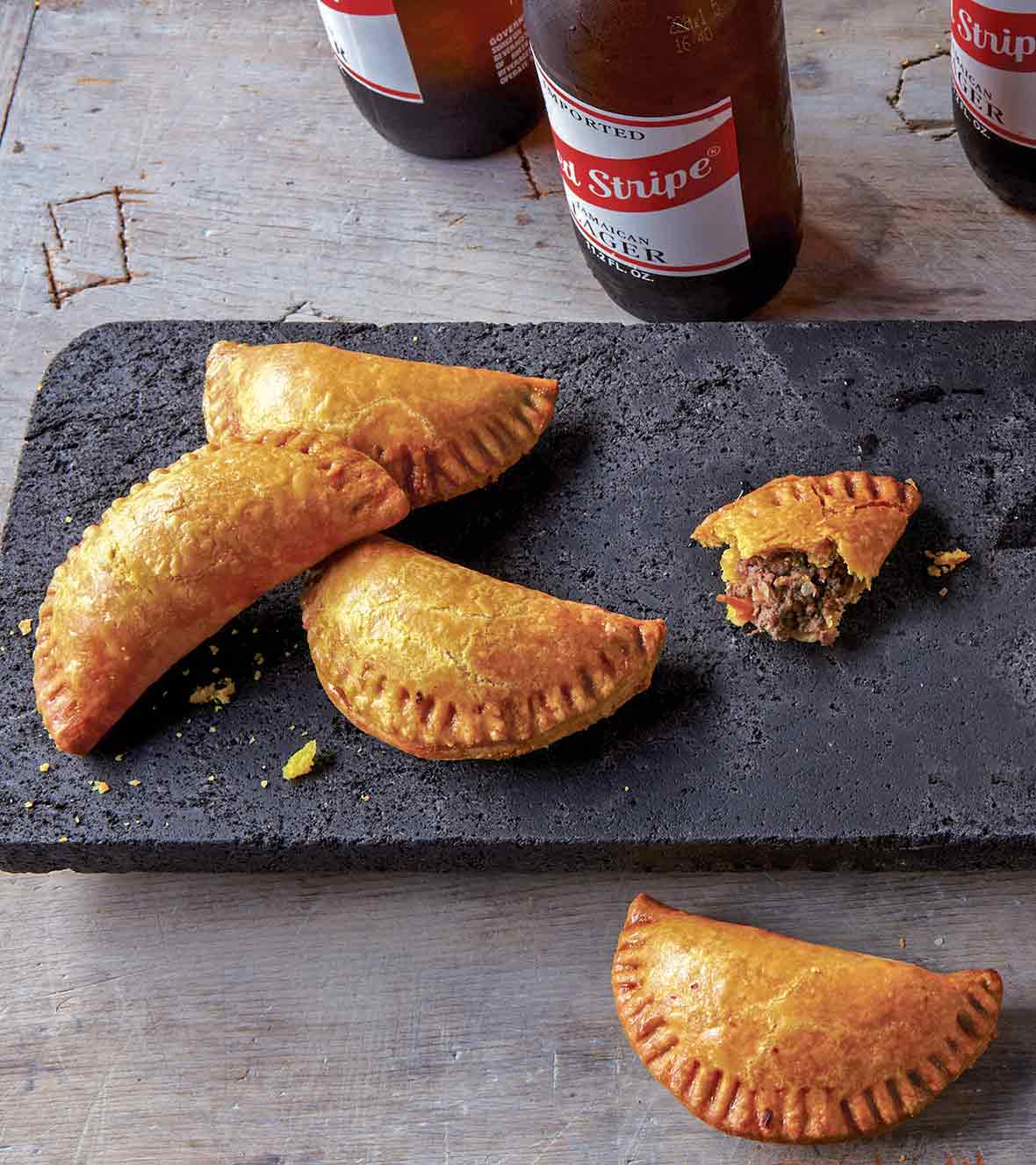 Five half-moon pastries, Jamaican beef turnovers, on a stone plate, three bottles of beer