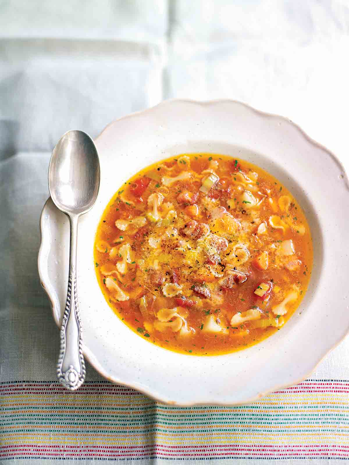 A white bowl filled with pasta soup with potatoes and pancetta on a linen cloth with a spoon resting on the edge of the bowl.