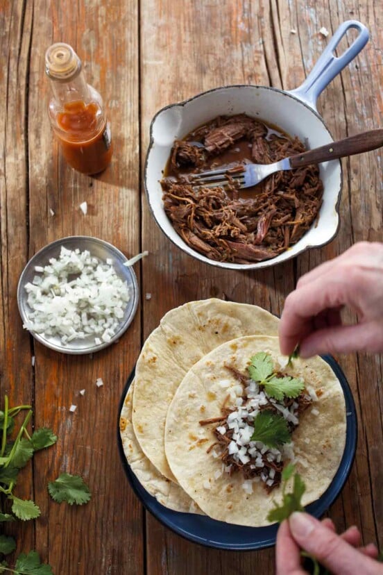 A person assembling a slow cooker barbacoa taco with a pot of shredded barbacoa meat, a small dish of onions, a bottle of hot sauce, and some cilantro around the taco plate.