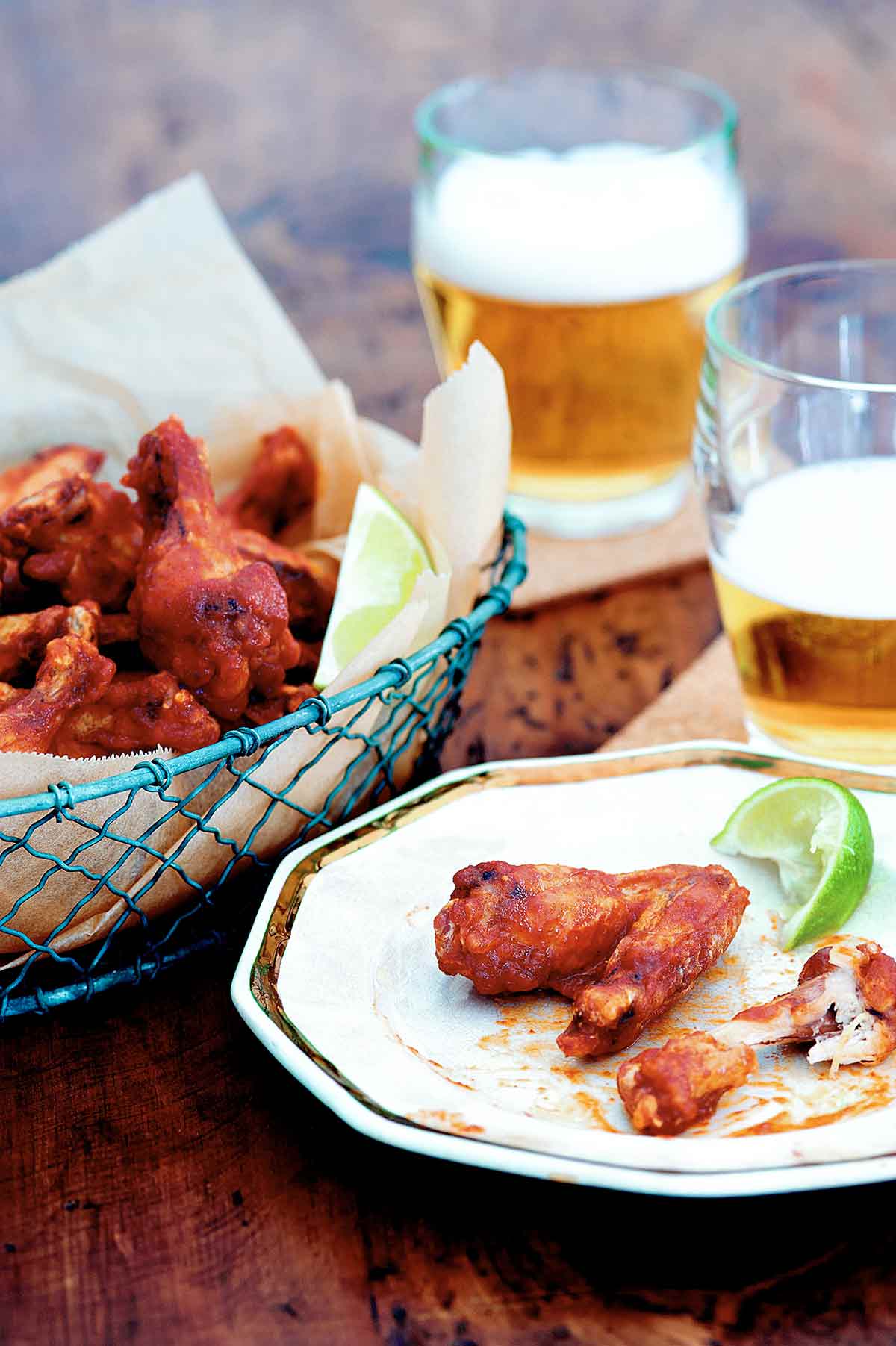 A basket of Sriracha chicken wings, two glasses of beer, and a plate with one eaten and one uneaten chicken wing and a wedge of lime.