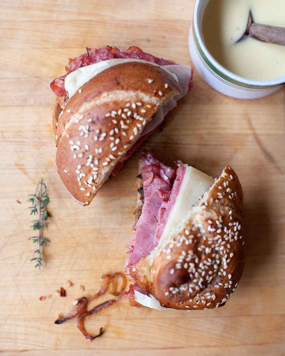 A pastrami, caramelized onion, and Gruyère sandwich on a pretzel roll sliced in half with a sprig of thyme lying beside it on a wooden cutting board.